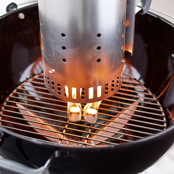 Ein silberner Weber Rapidfire Anzündkamin steht auf dem Grillrost eines schwarzen Weber Holzkohlegrills. Unter dem Anzündkamin brennen Anzündwürfel mit hellen Flammen, die durch die Löcher im Kamin sichtbar sind.